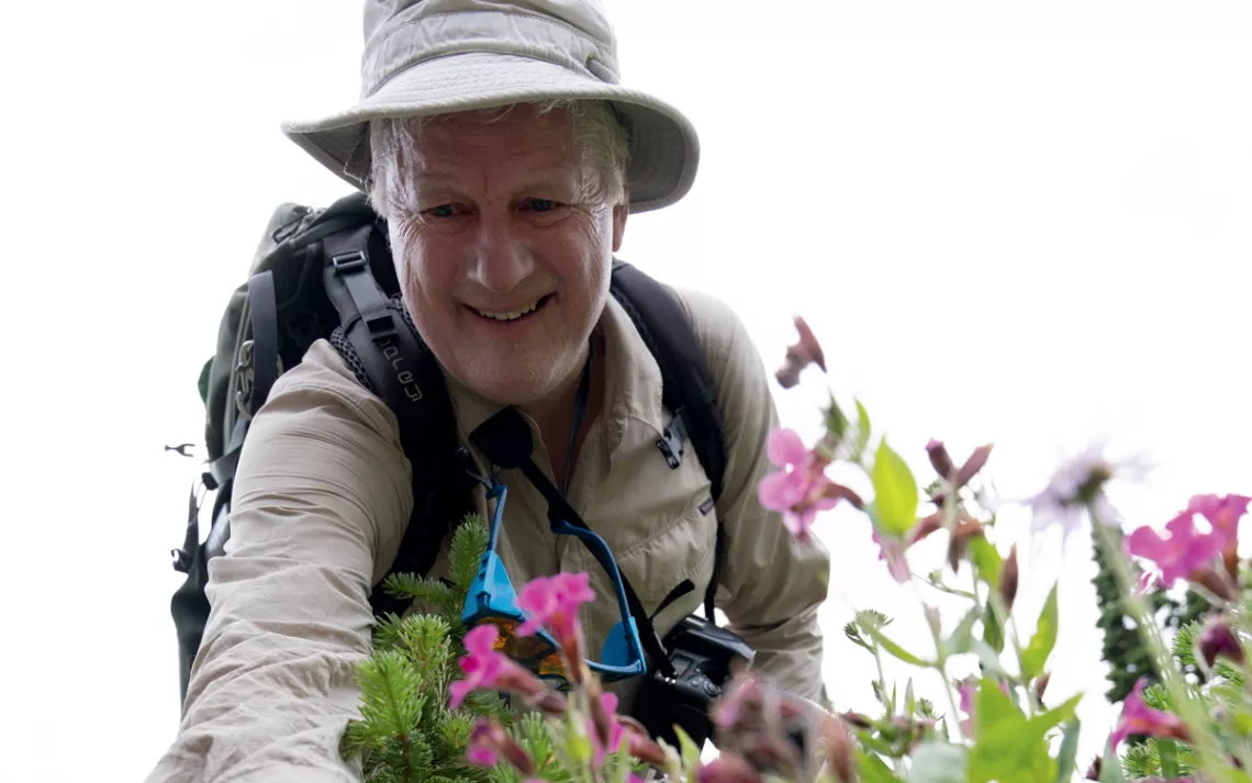 Harvey Locke, wearing a backpack and beige field hat, smiles and reaches his arm out toward some pink flowers.