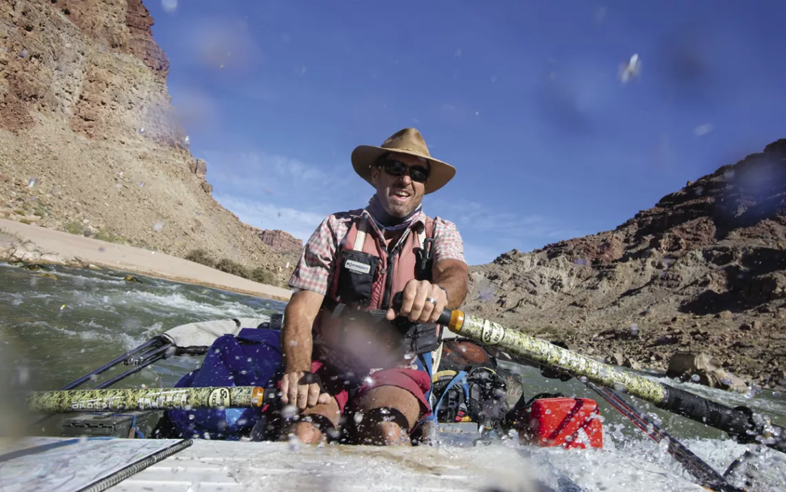A man steers a boat on a river with two oars. Behind him is blue sky and desert/rock walls.