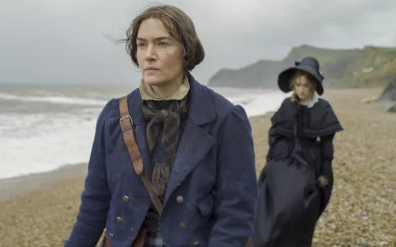 Kate Winselt and Saoirse Ronan walk along a blustery beach wearing bonnets looking grim