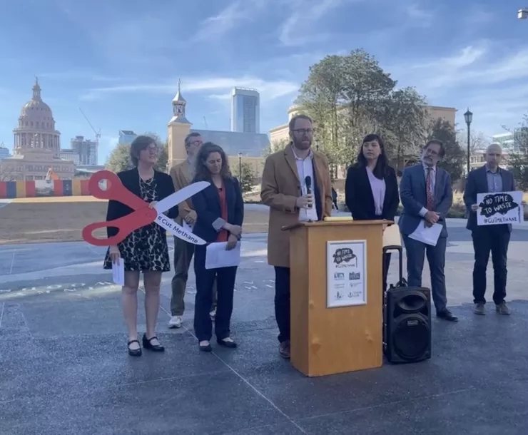 Seven people stand behind a podium with the Texas Capitol in the background. A woman holds a giant pair of scissors that say "Cut methane"