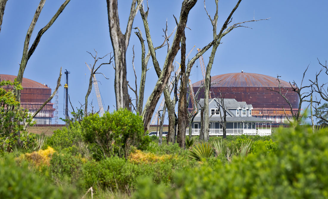 Gas storage tanks under construction seen through dead trees in a marsh, with a home in between.