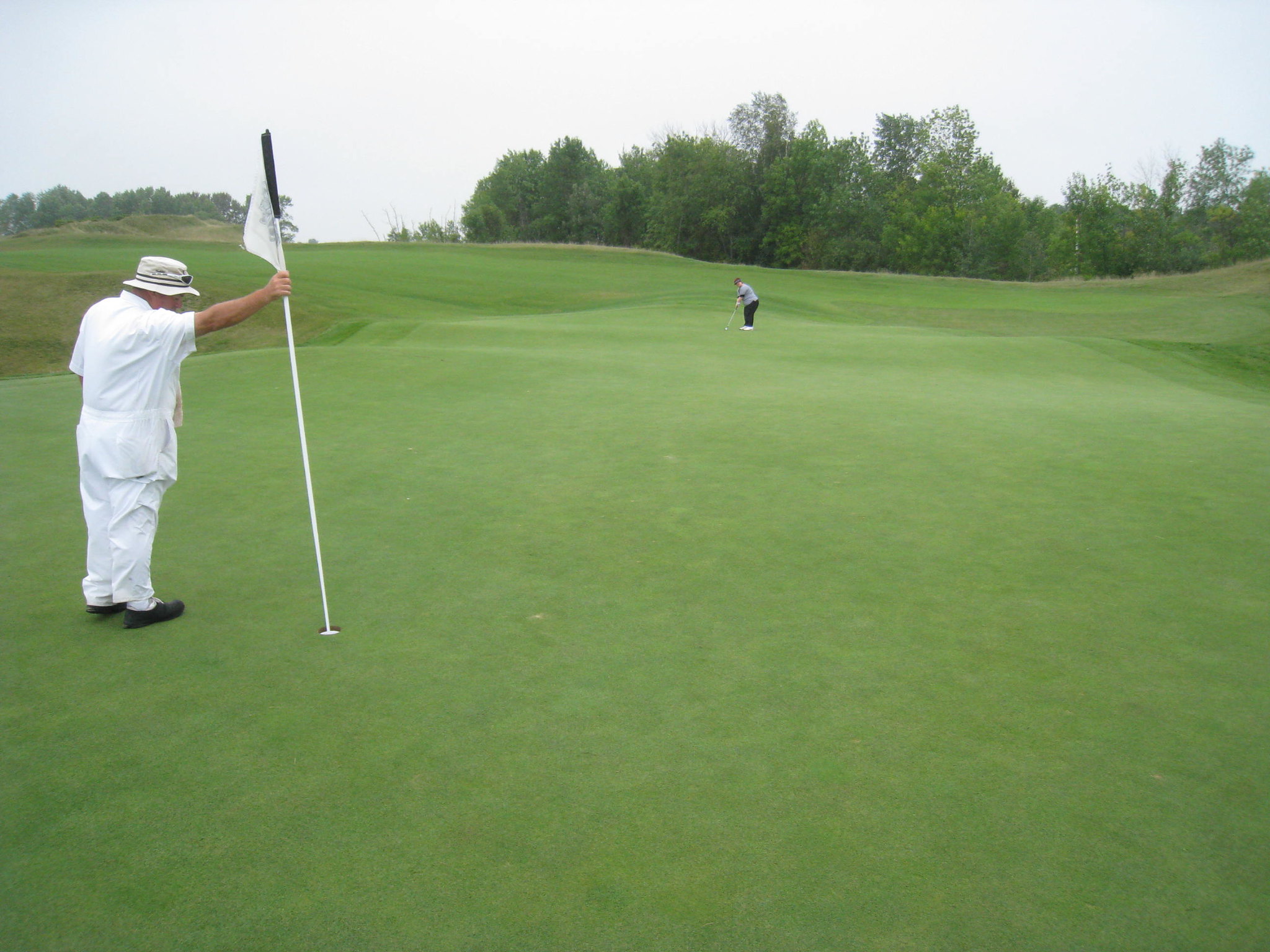 A man holds the flag on a golf course while a woman putts.