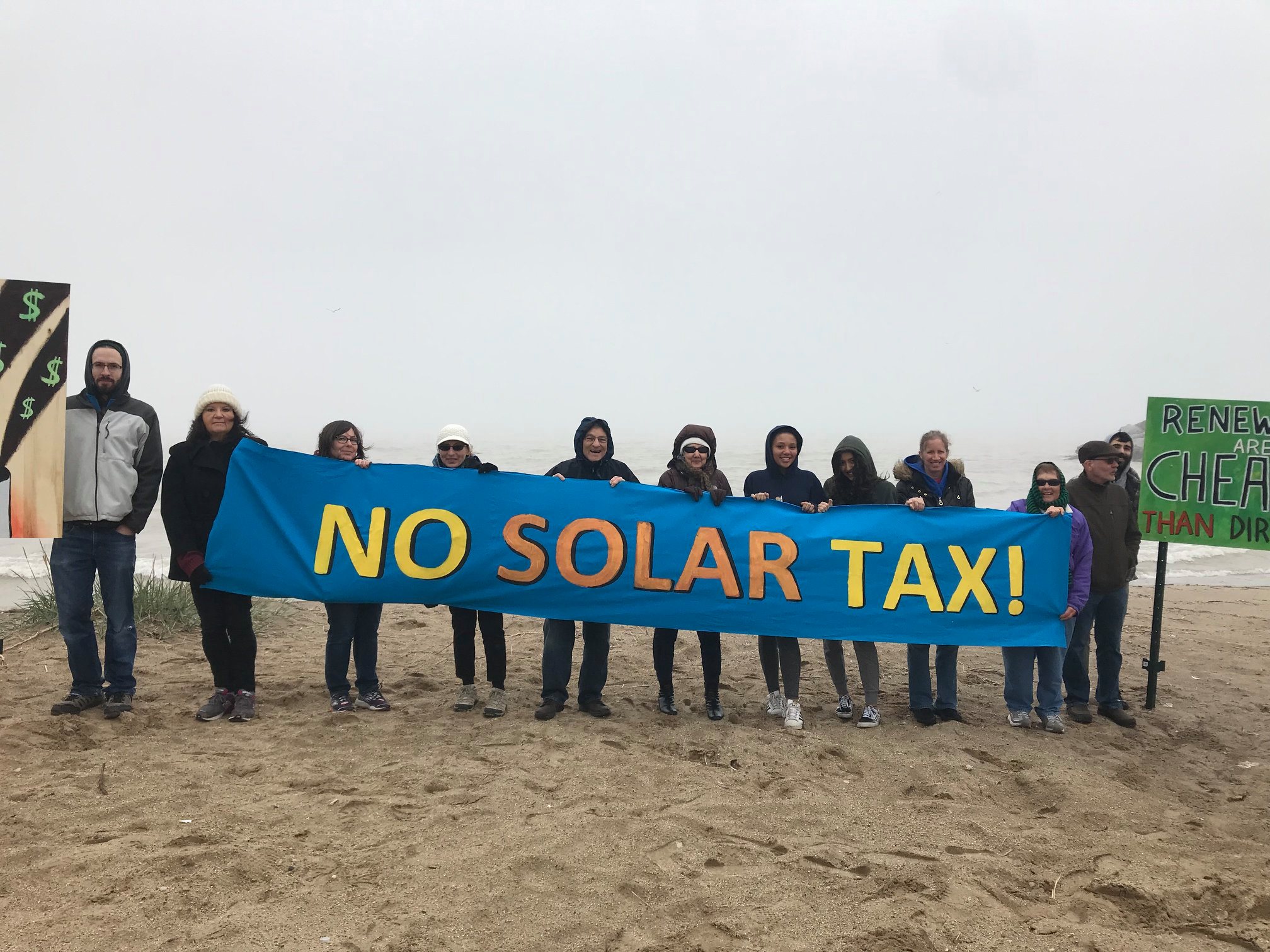 A group protesting at a beach holds a sign that reads "No Solar Tax!"