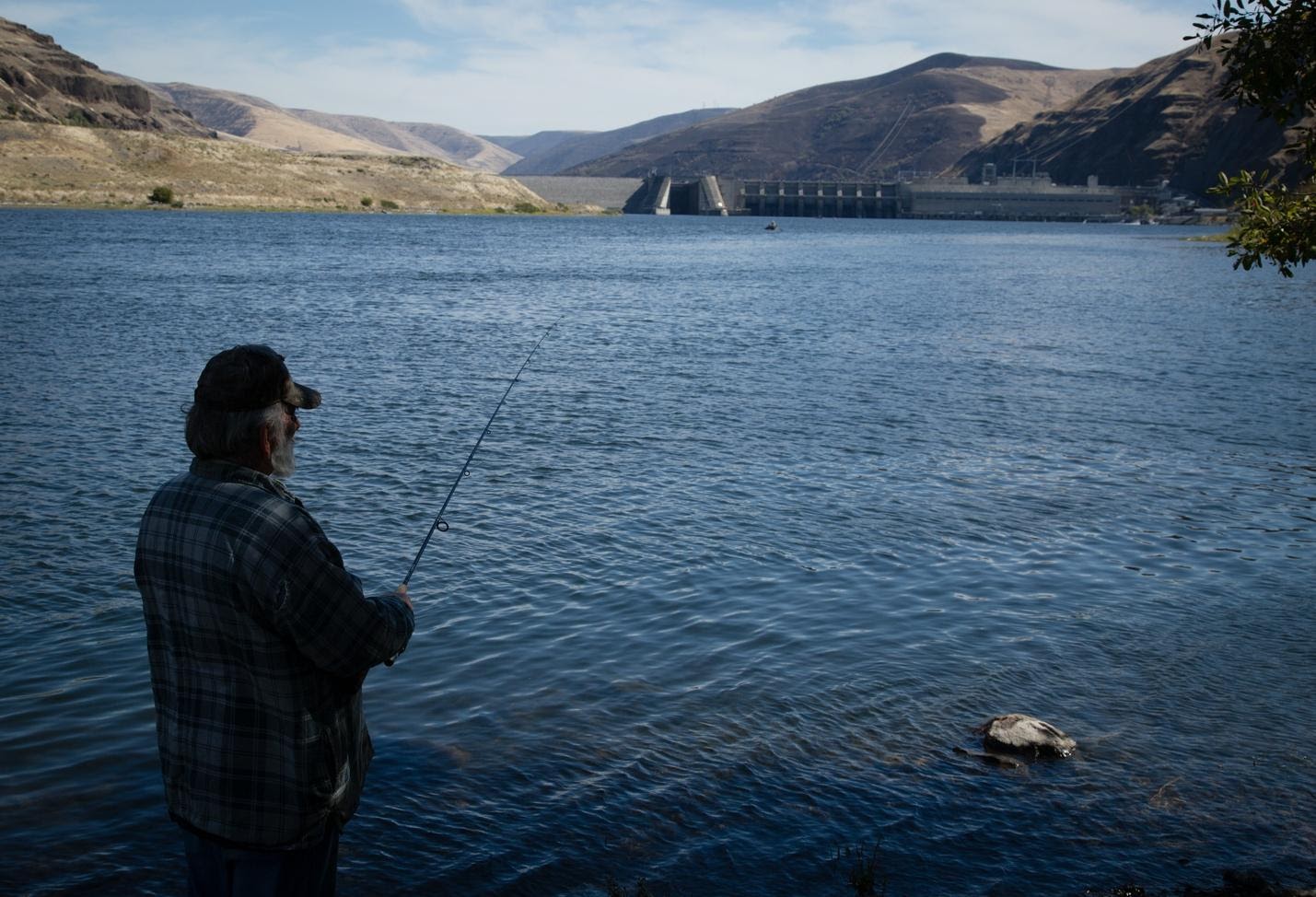 A man in a plaid flannel casts his fishing line below a dam on a wide blue river