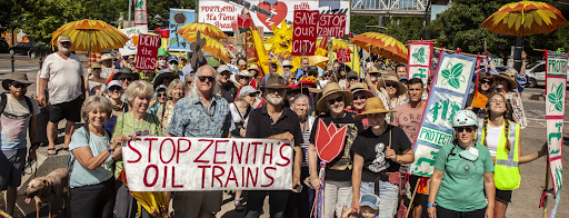 A crowd of 30+ activists gathered for a photograph on a sunny day behind a white banner that reads "Stop Zenith's Oil Trains" in red letters. Others hold signs that say "Deny the LUCS", "Save Our City", and "Stop Zenith", and sunflower umbrellas.