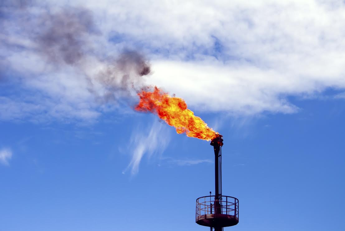A refinery against a dark blue sky. There is a huge plume of red and yellow fire erupting from a flare, spewing methane and fire into the air.