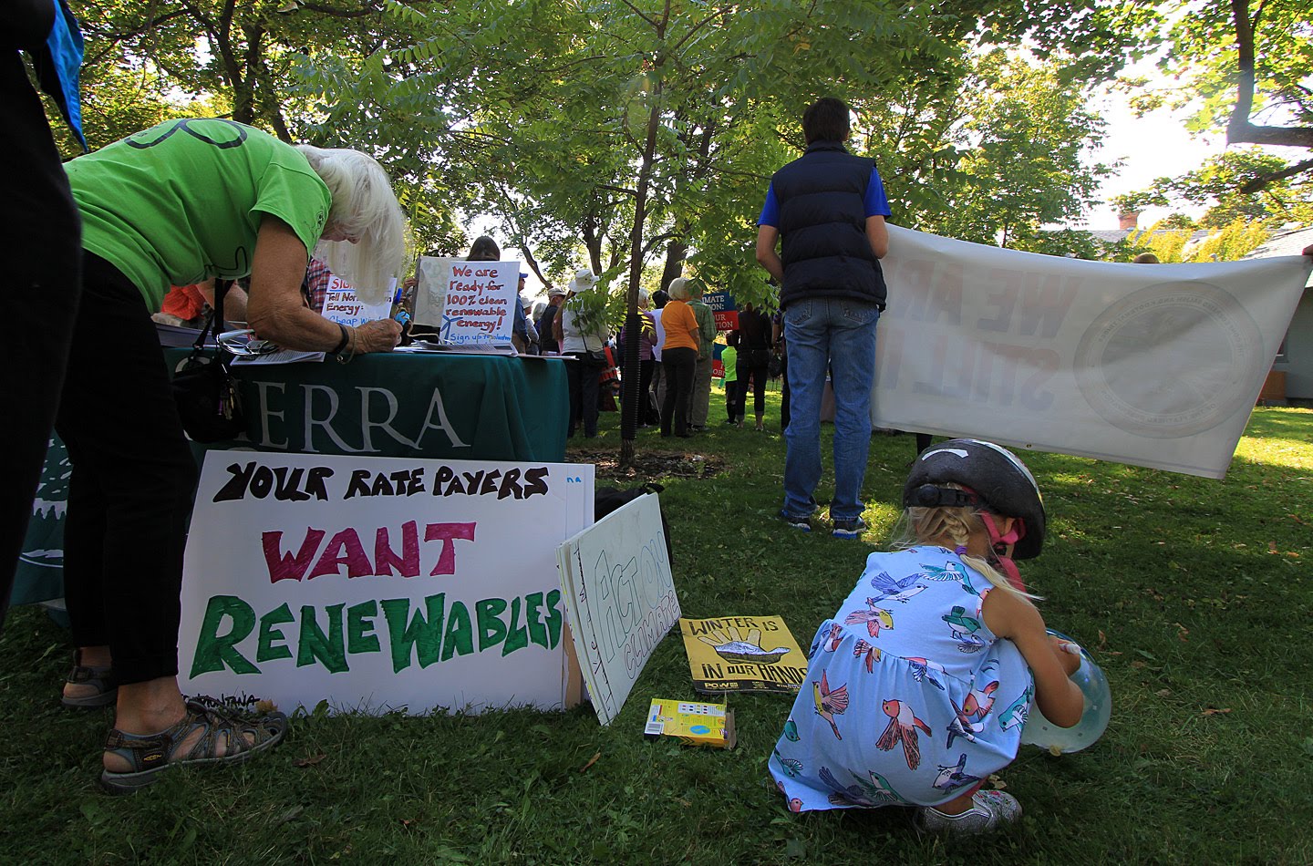 A small child crouches on the grass next to a sign reading "Your Ratepayers Want Renewables" while a group of people stand at a rally in the background.