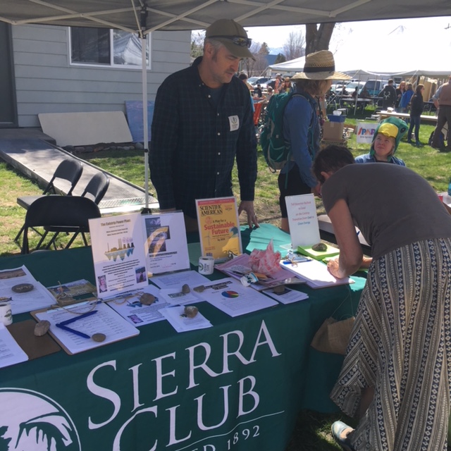 A volunteer stands behind a table draped with a Sierra Club banner while someone signs a piece of paper on the table.