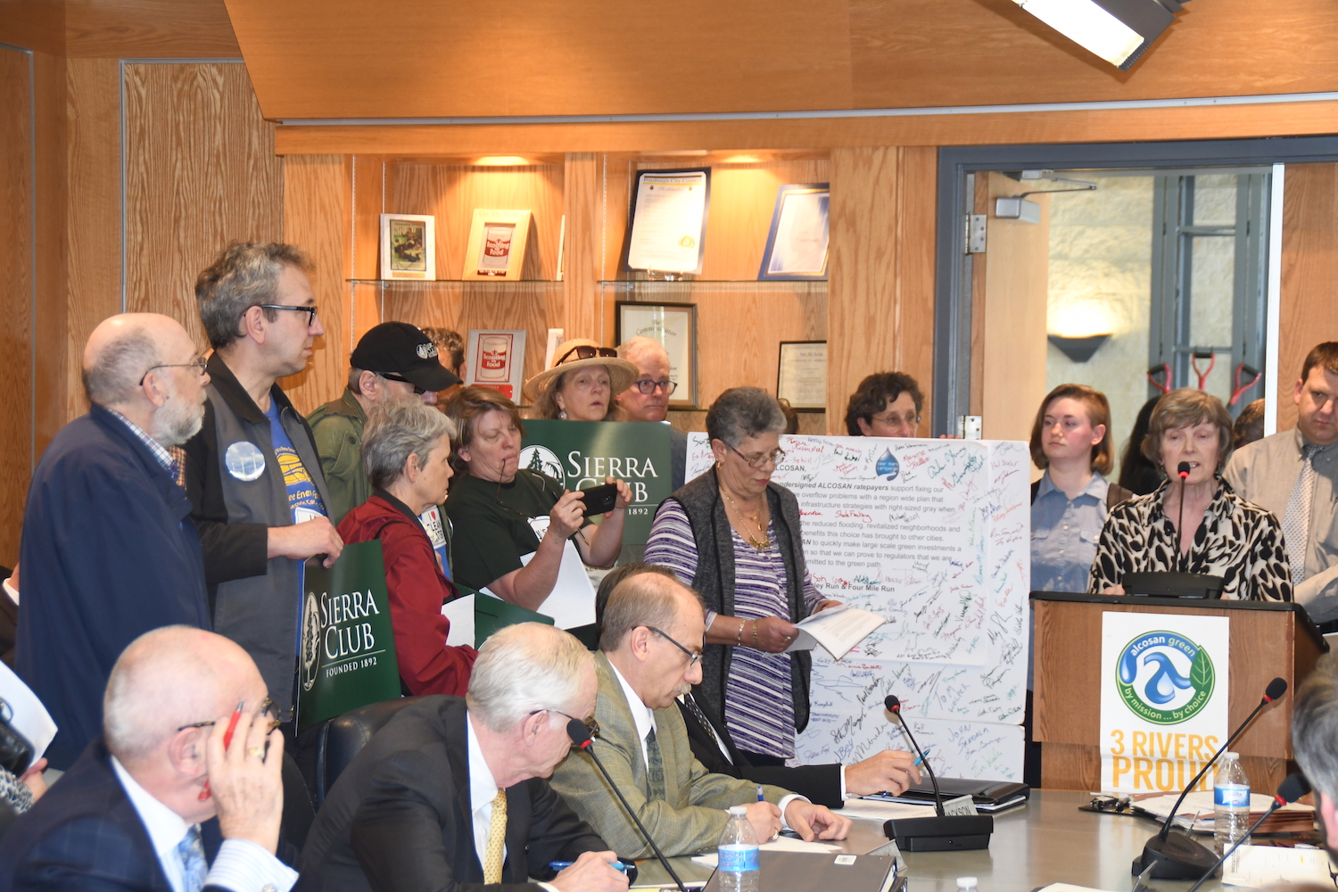 A dozen Clean Rivers Campaign supporters holding Sierra Club signs and a four-foot letter with countless signatures gather around a podium while a resident speaks out for clean, green investments. Three men in suits sit at a conference table in front of the podium.