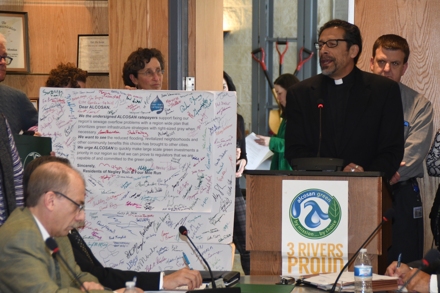 A man in a black suite wearing a white clerical collar and glasses speaks at a podium in support of green sewer infrastructure investments. Other supports stand nearby and a bald man in glasses sits at a conference table in front of the podium.