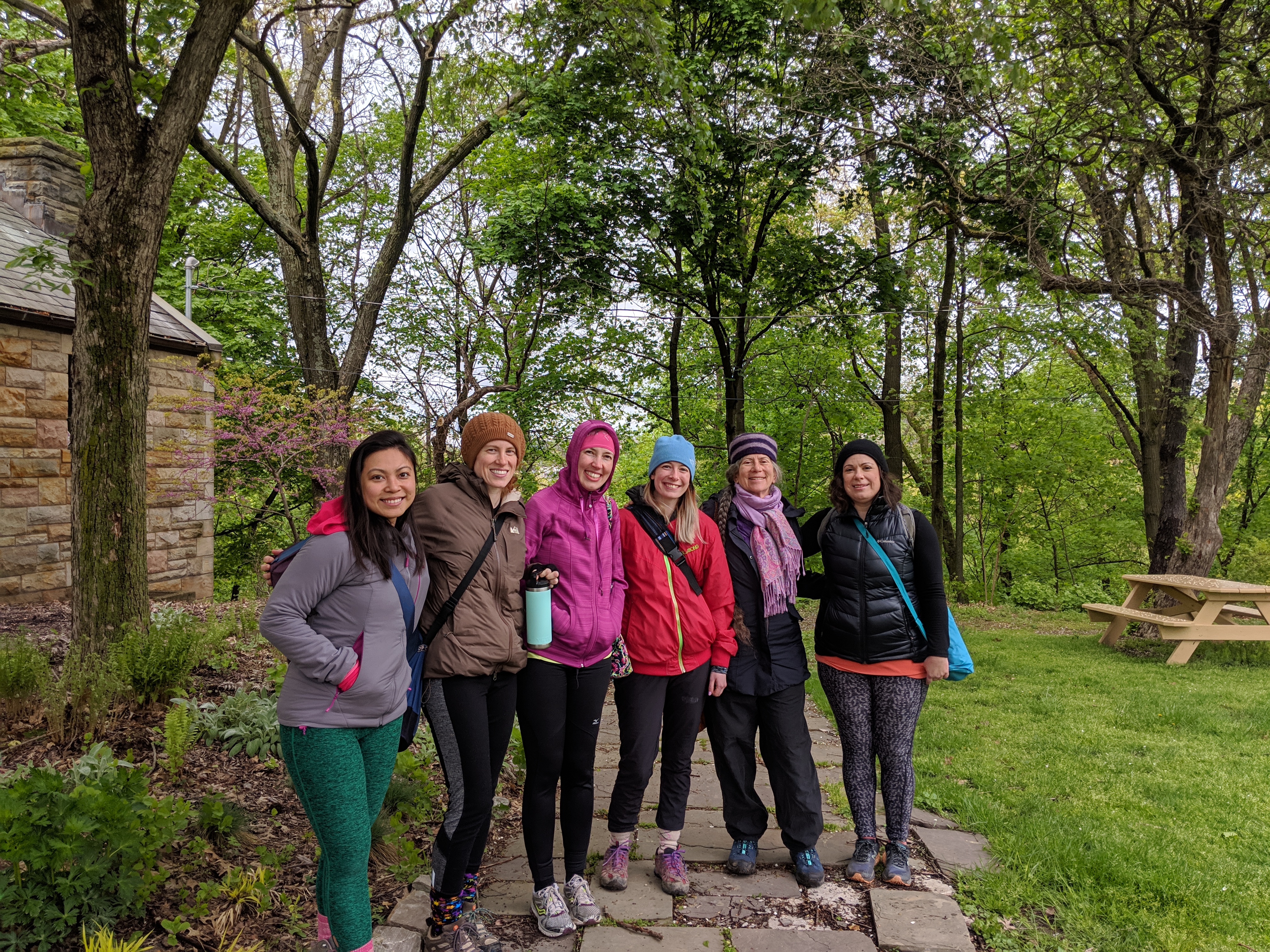 Six people wearing colorful spring jackets gather for a group photo outside a stone building with a cherry blossom in the background.