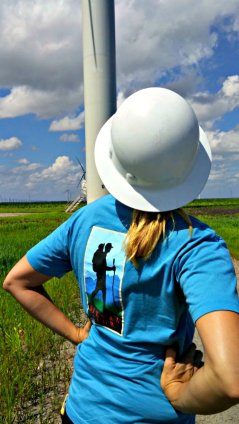 A person in a Sierra Club t-shirt and a hard hat stands in front of a wind turbine, looking up at the blades