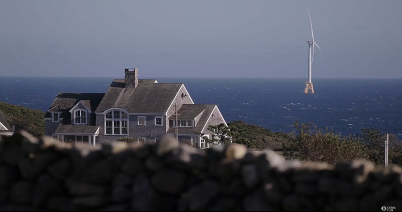 A wind energy turbine off Block Island, RI, is shown from the island, with a large home and stone wall in the foreground