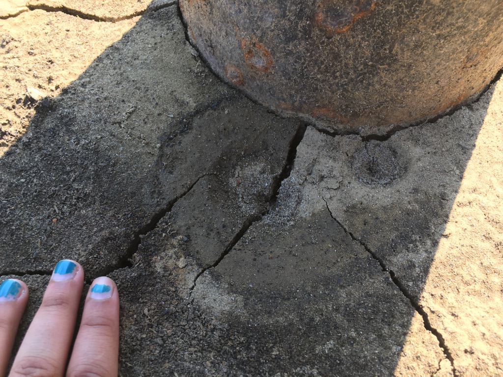 A brown hand with chipped, teal, nail polish is placed on cracks in the ground next to an orphan well, likely the result of escaping gas.
