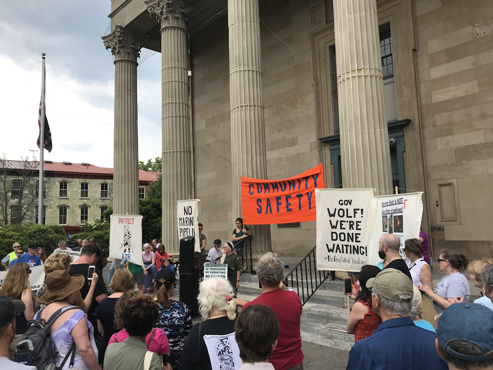 A group of residents gather on the steps of the West Chester Historic Courthouse to demand a halt to the Mariner East 2 Pipeline. An impacted landowner, Ellen Gerhart, is at the podium speaking to her experiences. People in the crowd hold signs that say "COMMUNITY SAFETY" and "Gov. Wolf! We're done waiting!"