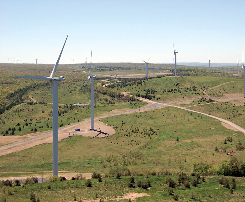 Five wind turbines stand on a hilly landscape with half a dozen wind turbines in the distance