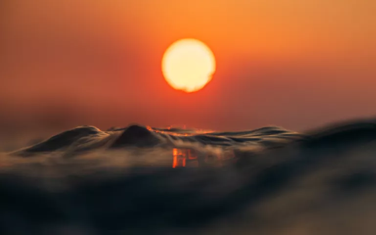 A water-level view of small waves and the sun against an orange sky