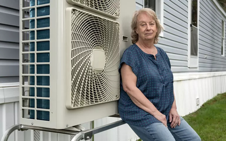  Anne Pappas sits next to her heat pump, installed on the side of her gray and white mobile home.