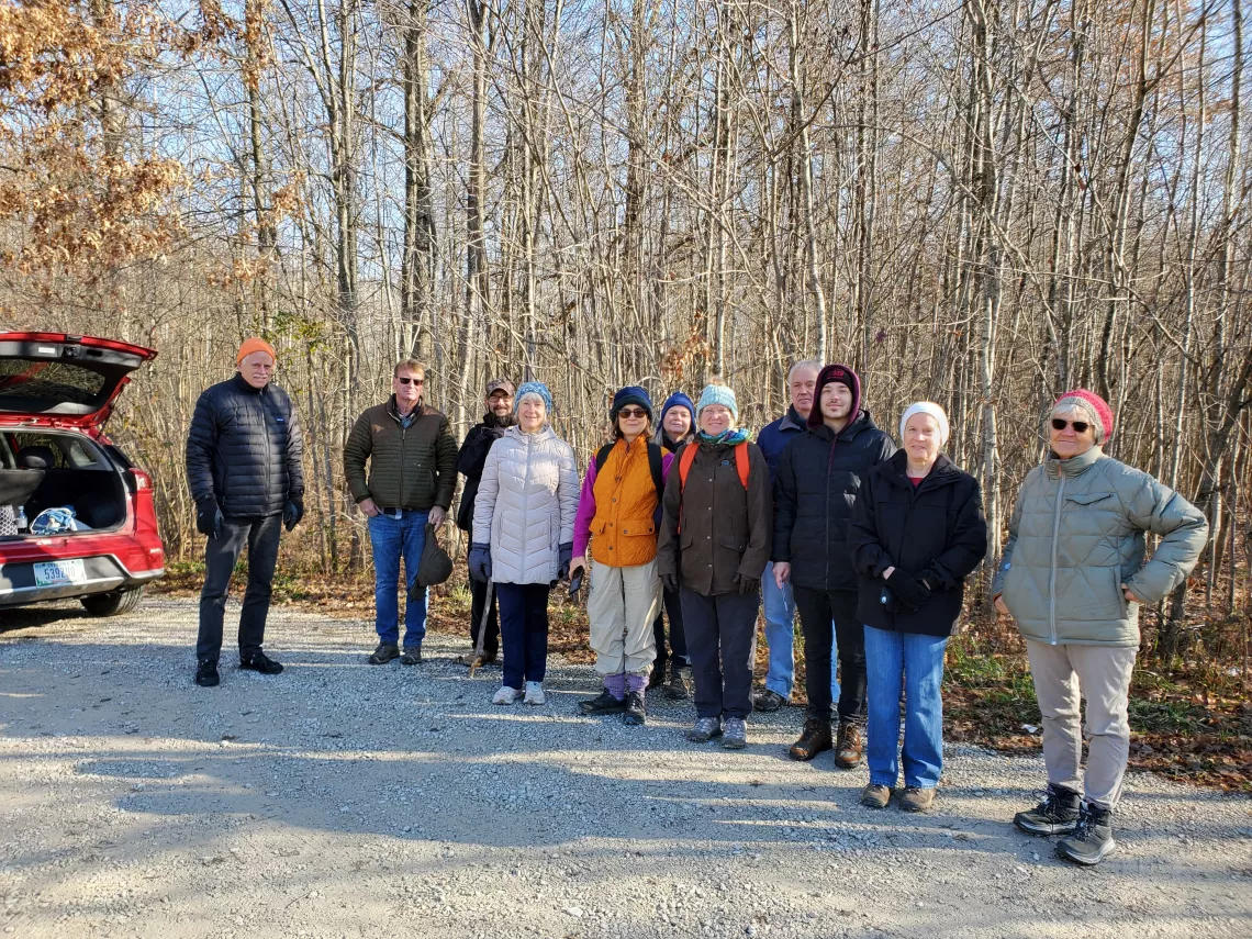 A group of people stand in a parking lot at a trail head of a forest, about to go on a hike. They are dressed for the cold weather and smiling.