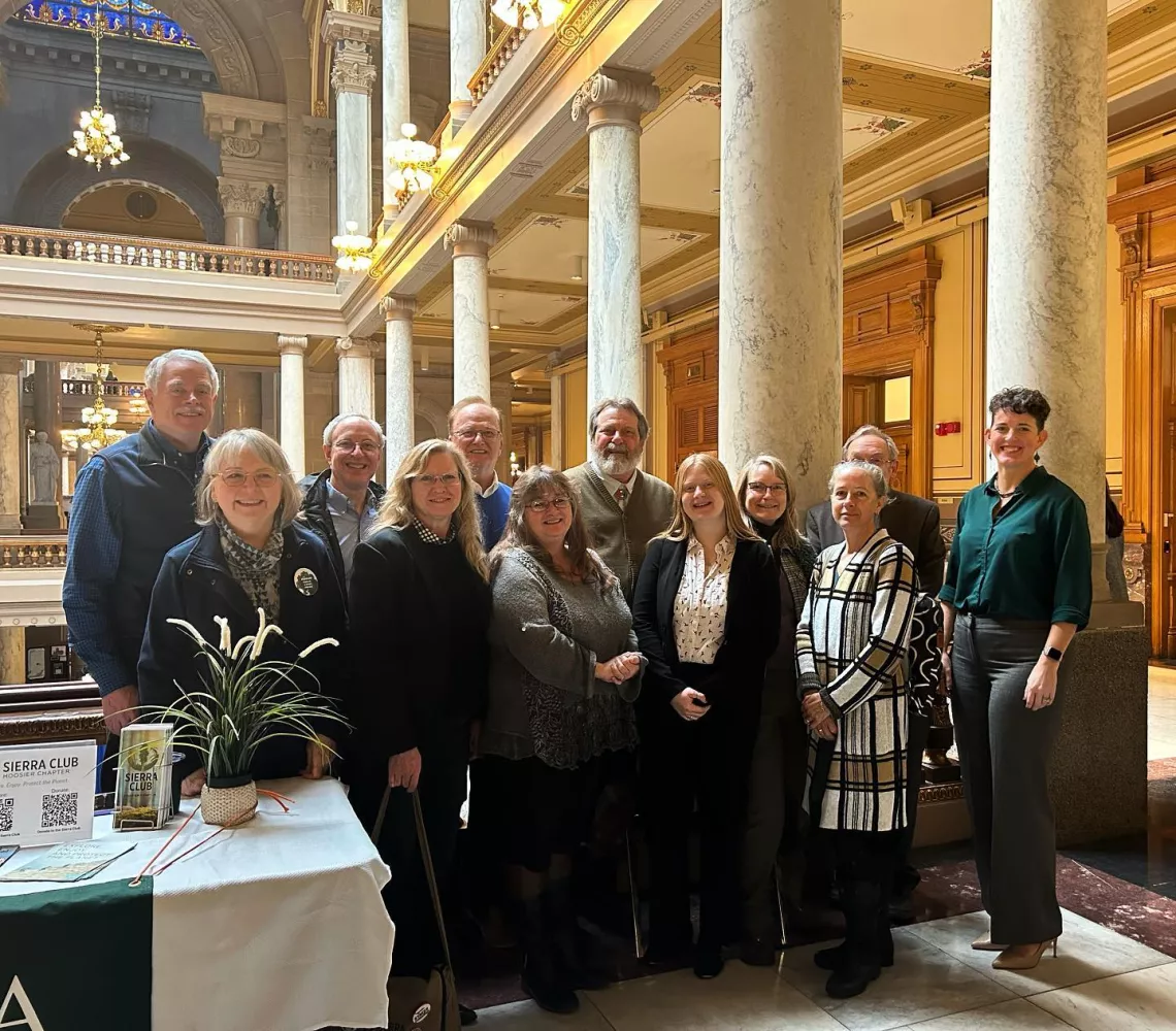 A group of 12 people standing for a photograph at the Indiana Statehouse. To the left, half of a table is in the photo which shows the Sierra Club logo.