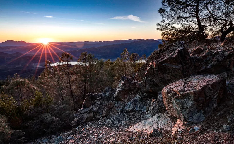 Image of the rocky ridge of Molok Luyuk with a view of trees below and sun rising/setting among the mountains in the distance