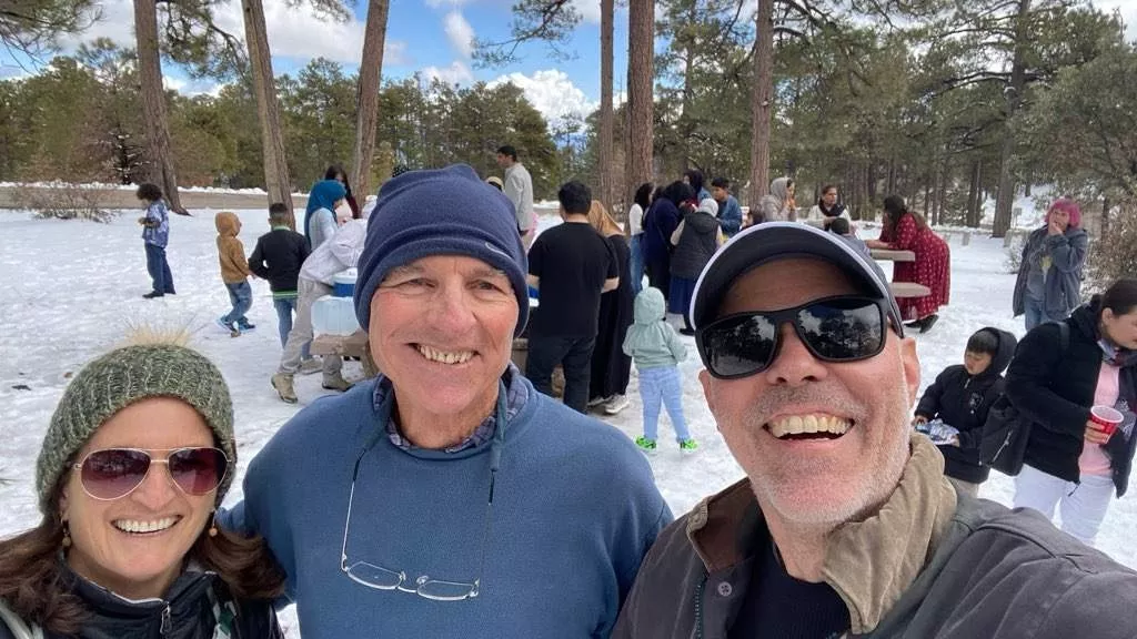 Three people wearing warm hats, smiling at the camera while people play in the snow behind them.