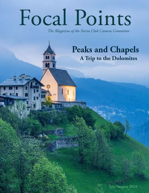 A tall white chapel with a slate roof is perched atop a green mountain with pearly blue sky in the background