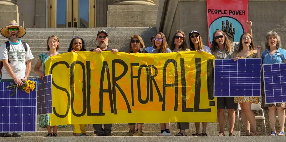 Picture of people holding a big yellow banner that reads "Solar For All" with some people holding solar panel signs