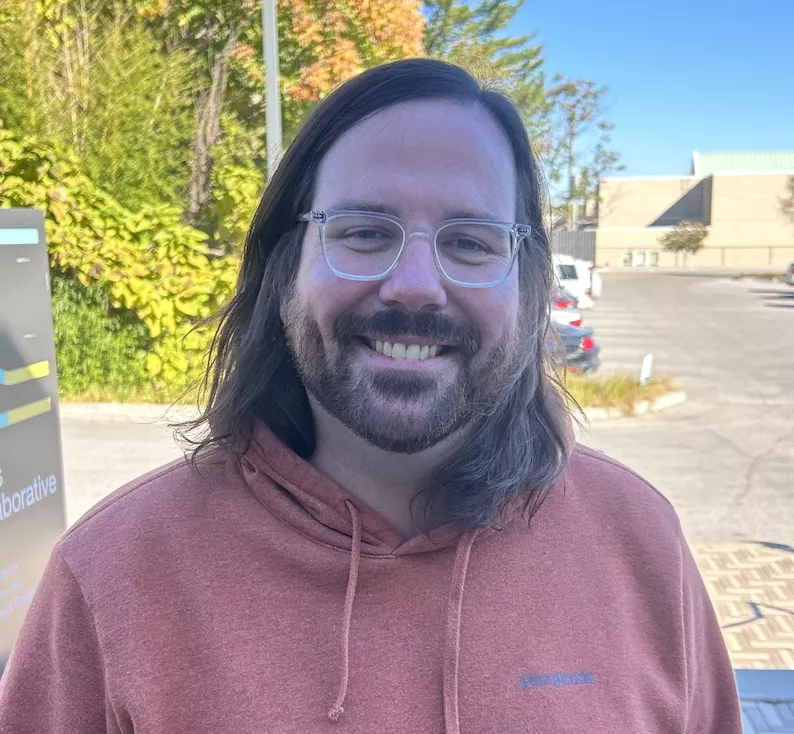 Ed is a white man with shoulder length brown hair and facial hair. He is wearing clear glasses frame and an organe hoodie. 