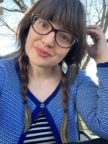 A woman with long brown hair in two plaits. She is wearing dark-rimmed glasses and a blue and white cardigan. There is a blue sky and a bare tree just visible behind her. She is smiling.