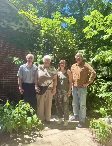 Four people stand in a garden. The leaves on the trees are green and blue sky can be seen beyond the trees. The people are smiling and looking at the camera for the photo.