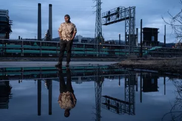 Qiyam Ansari stands in front of industry and railroad infrastructure with his reflection mirrored in a body of water.