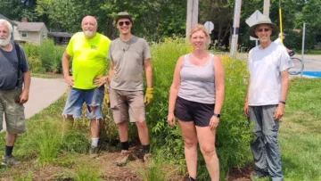 Five people stood by a path with pollinator plants behind them. It's a warm day and most are wearing shorts. They're smiling for the photo and ready to get to work maintaining the path.