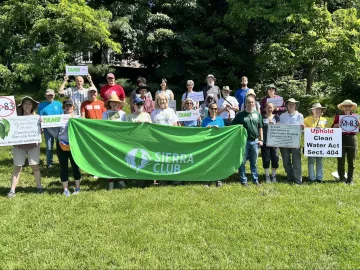 Image of group of volunteers holding up green Sierra Club banner, TAME coalition signs, and "no M83 highway" signs, on a lush green grass field with tress in the background. 