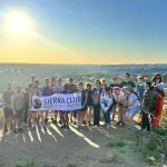 Picture of lots of people standing at top of a mountain overlooking a city holding a banner that says "Sierra Club OUTdoors Idaho" with the sun setting