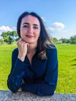 A thin, white woman with medium-length brown hair rests her chin on her hand and smiles against the backdrop of a lush green park.