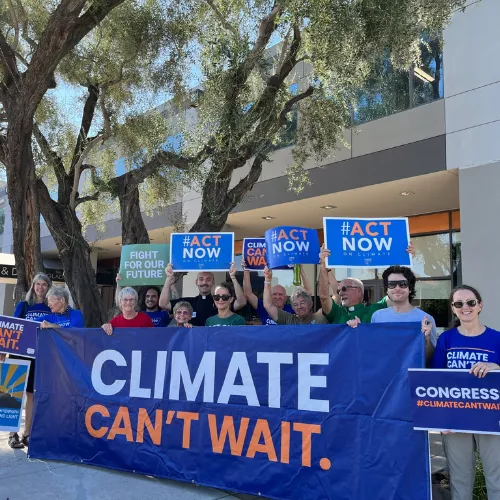 A small crowd gathered in front of a building with signs that say "Climate Can't Wait"