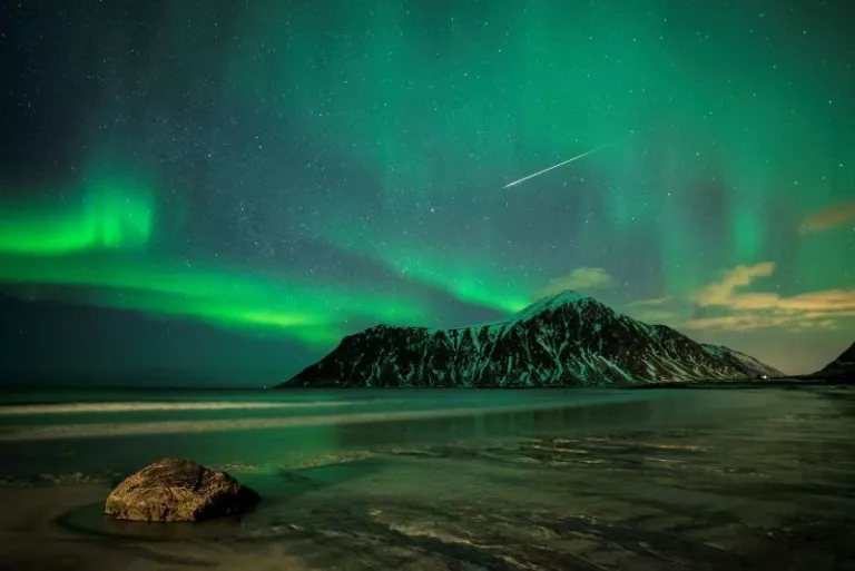 Aurora Borealis lights up the sky in green ribbons and a comet draws a white line across the sky. The foreground is a froze shore that reflects the green in the sky.