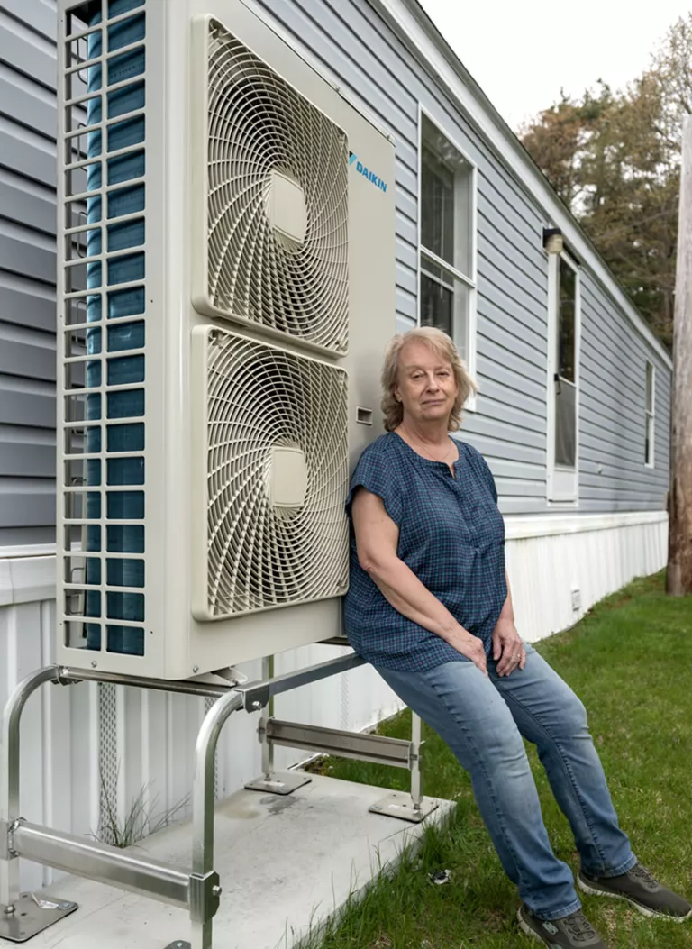   Anne Pappas est assise à côté de sa thermopompe, installée sur le côté de son mobil-home gris et blanc.