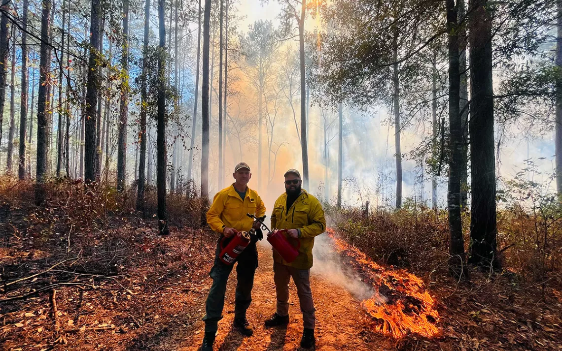 Two men wearing yellow PPE and holding ignition canisters, and standing amidst a ring of fire during a prescribed burn in Southwestern Georgia