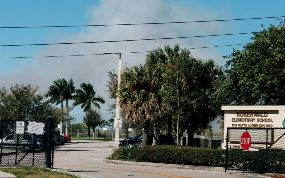 A road goes past a sign for Rosenwald Elementary School with smoke plumes in the distance