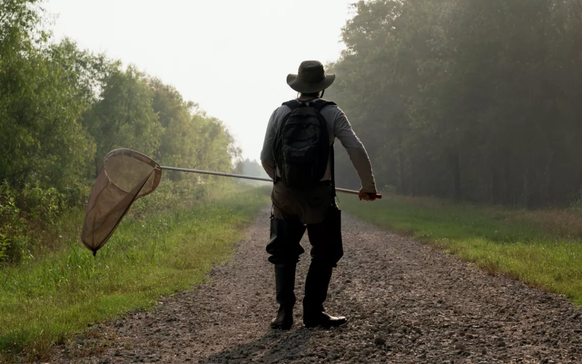 Jim Childress from behind on a rocky trail, wearing waders and a backpack and carrying a net