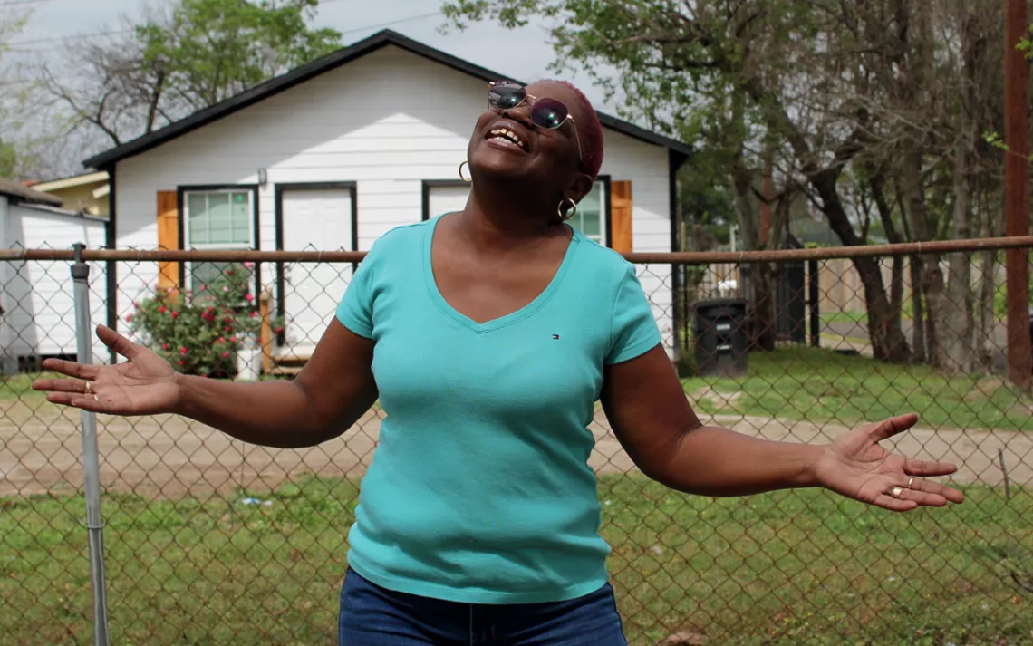 Sandra Edwards wears a turquoise shirt and sunglasses and holds her hands up to the sky and smiles. Behind her is a chain-link fence and a small white house.
