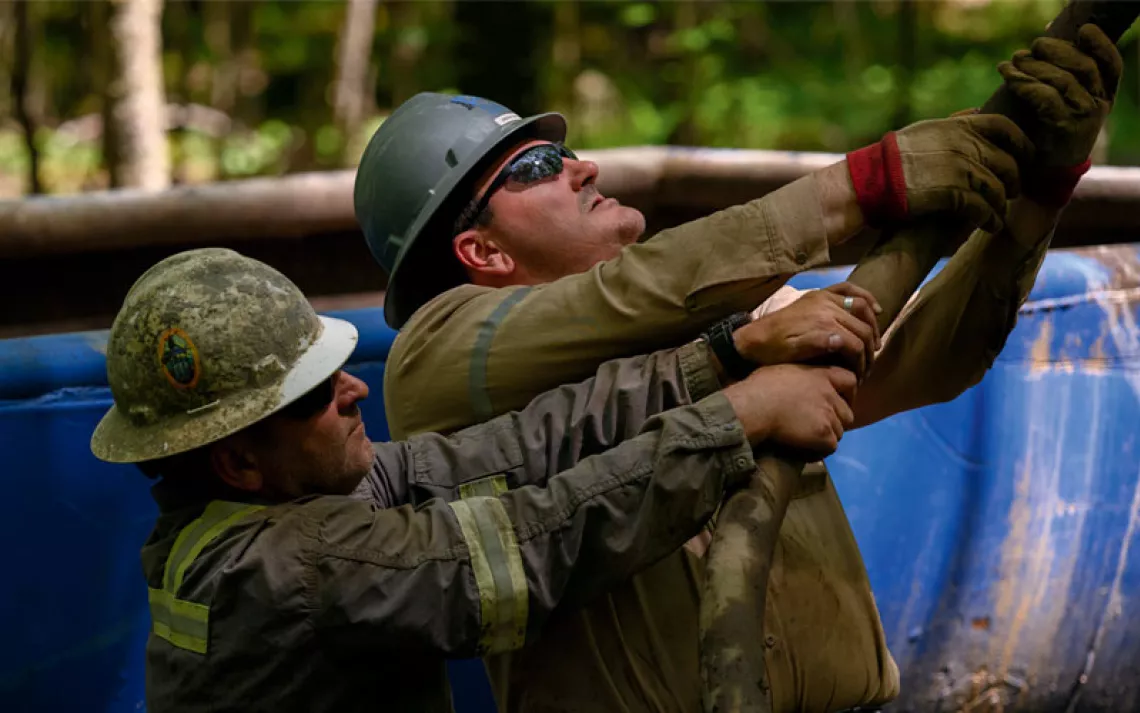 Two men in hardhats hold onto a cable at an abandoned oil well.