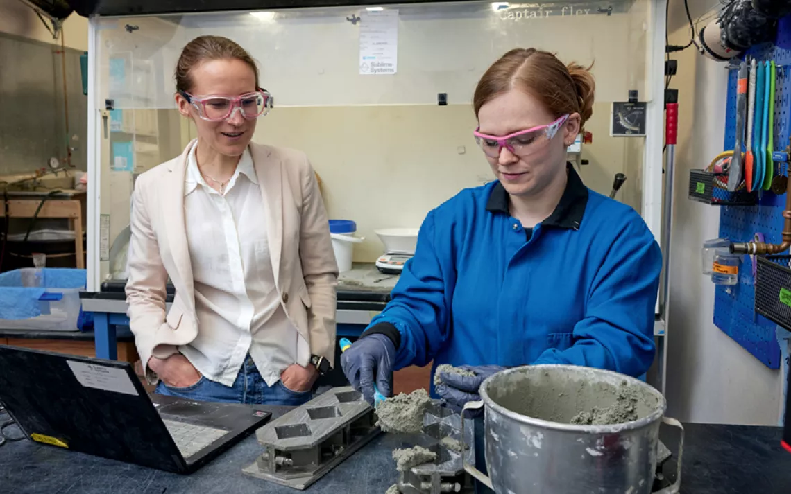 Leah Ellis wears a light blazer and white button-down shirt, watching Summer Camerlo-Bass in a blue work shirt scooping gray cement from a metal mixing bowl.