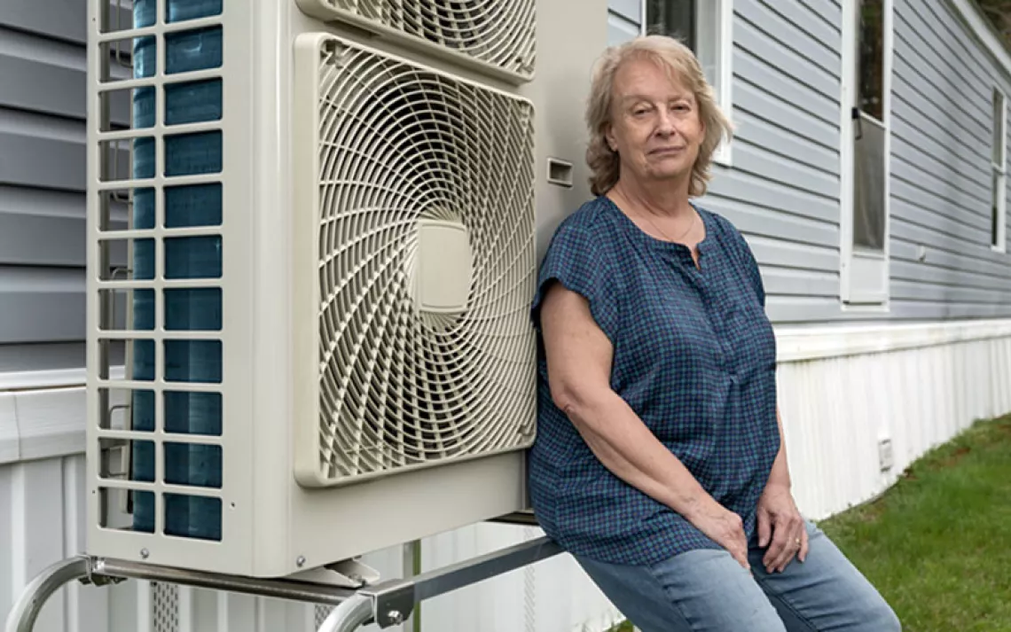   Anne Pappas est assise à côté de sa pompe à chaleur, installée sur le côté de son mobil-home gris et blanc.