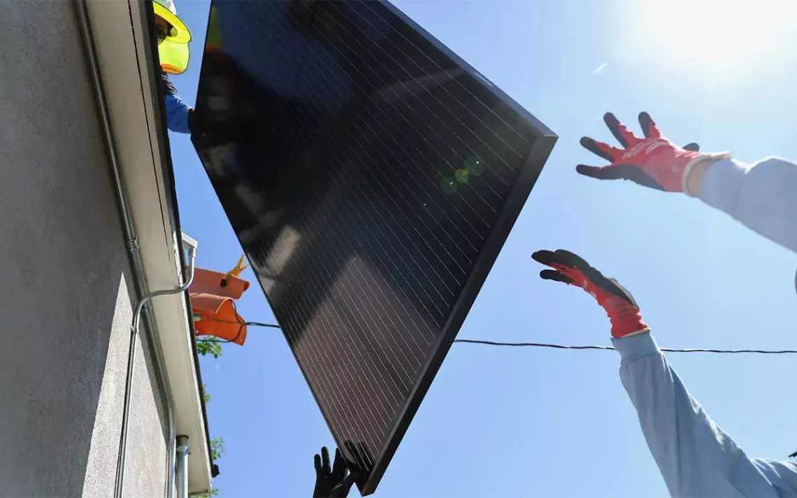 A worker hands off a solar panel being installed by another worker