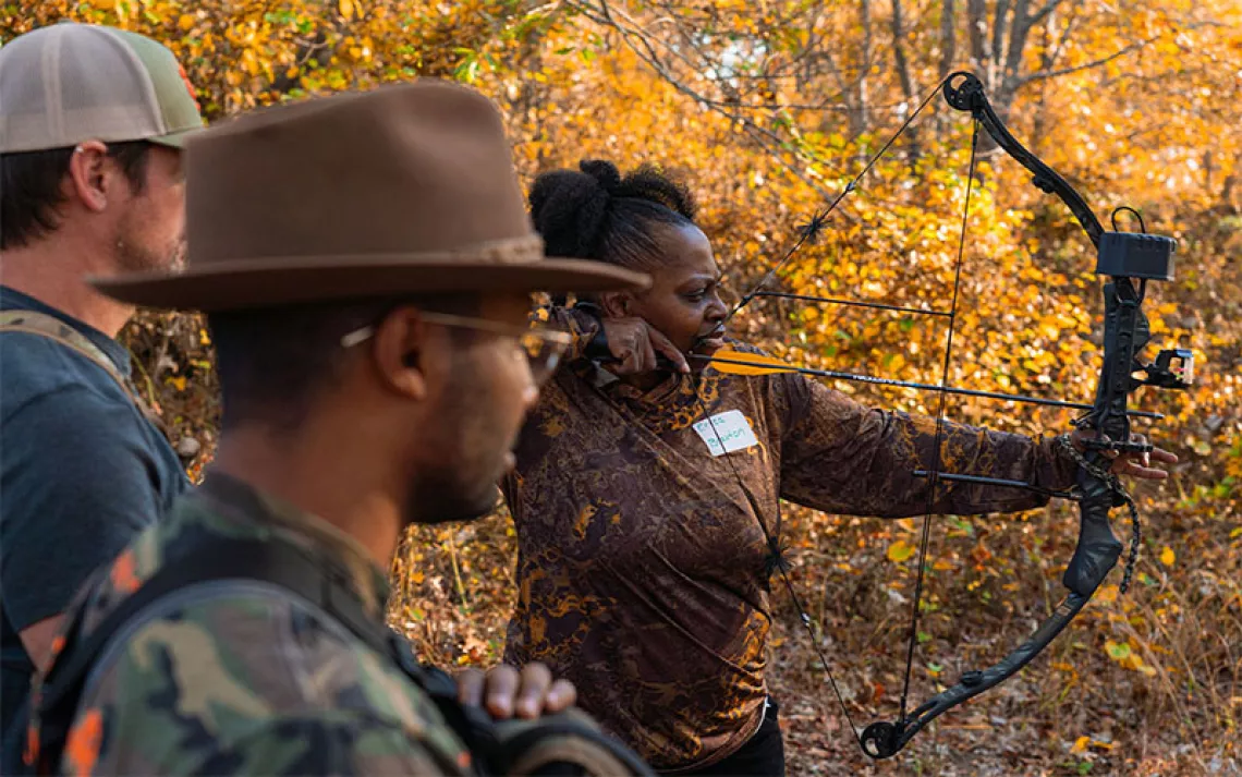 A Black woman in camo shoots a bow and arrow among fall foliage