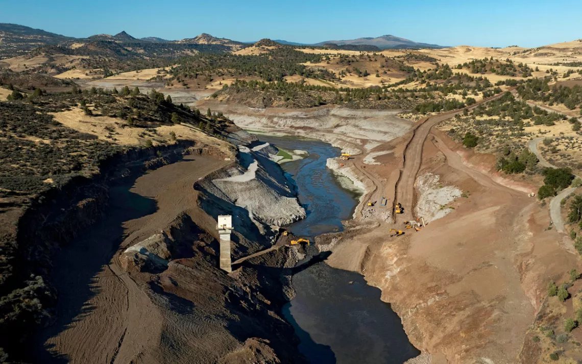 Aerial photo of the small cofferdam at the Iron Gate Dam being breached by an excavator as water flows through