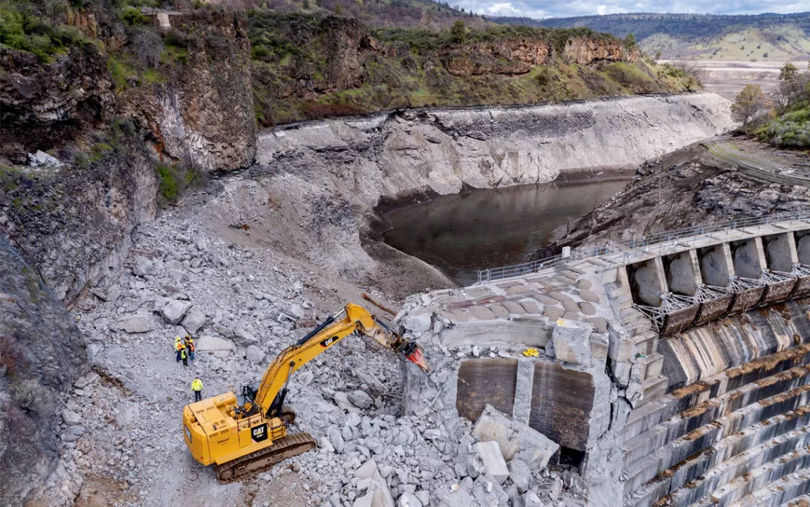 Aerial photo of the dismantling of Copco 1 Dam shows the water line of the former reservoir, an excavator, and workers.